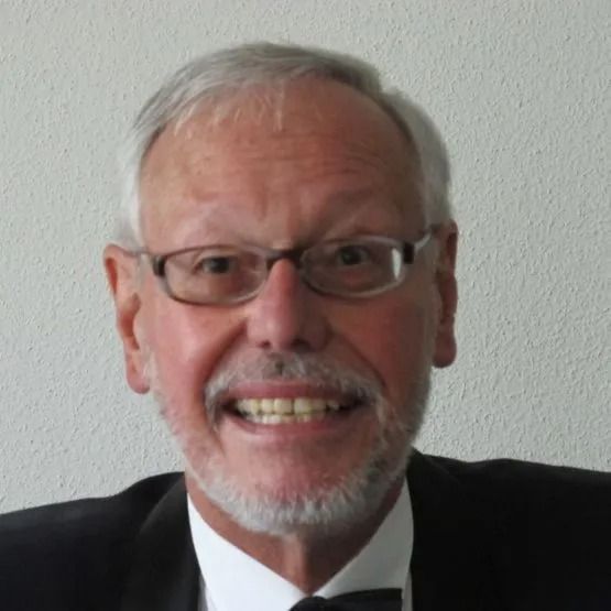Eine lächelnde Person mit grauem Haar und Bart, die eine Brille und einen formellen Anzug mit Fliege trägt, vor einer schlichten Wand.