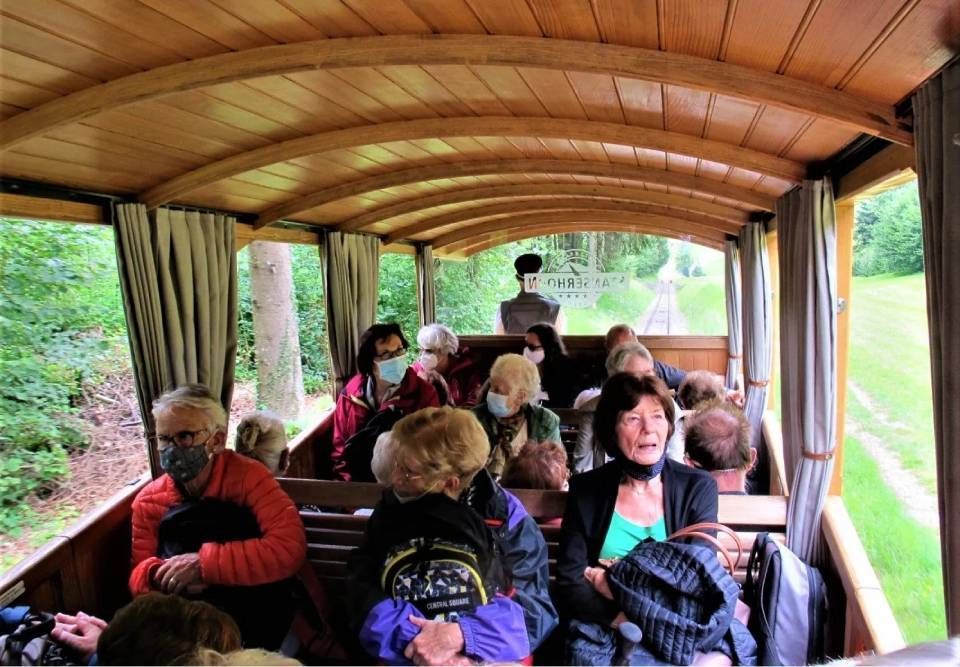 Eine Gruppe von Menschen mit Gesichtsmasken sitzt in einem offenen Holzzugwaggon, der durch einen grünen Wald fährt.