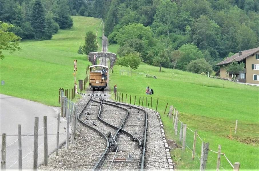 Eine Standseilbahn fährt auf Schienen durch eine grüne, hügelige Landschaft neben einer Strasse und einem Holzhaus.