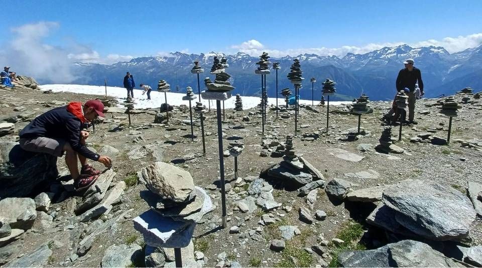 Menschen interagieren mit auf Metallstangen montierten, ausbalancierten Steinhaufen auf einem felsigen Berggipfel vor schneebedecktem Hintergrund.