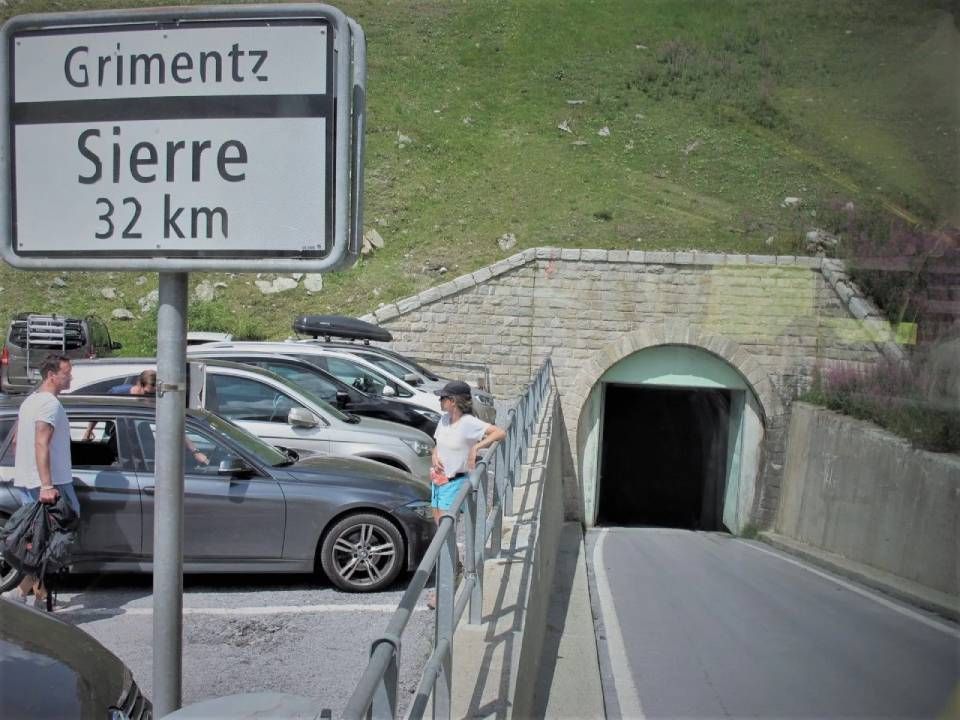 In der Nähe eines geparkten Autos und eines in einen grasbewachsenen Hang gebauten Tunneleingangs steht ein Strassenschild mit der Aufschrift „Grimentz“ und „Sierre 32 km“.