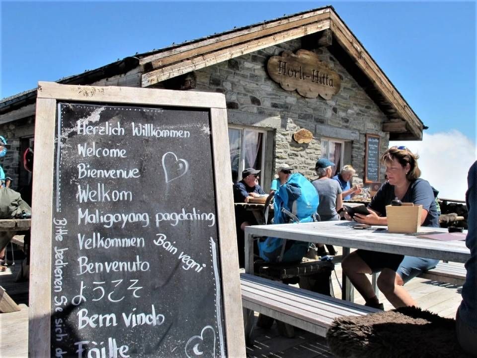 Vor einer rustikalen Berghütte aus Stein mit Sitzgelegenheiten im Freien prangt ein Kreideschild mit der Aufschrift „Willkommen“ in vielen Sprachen.