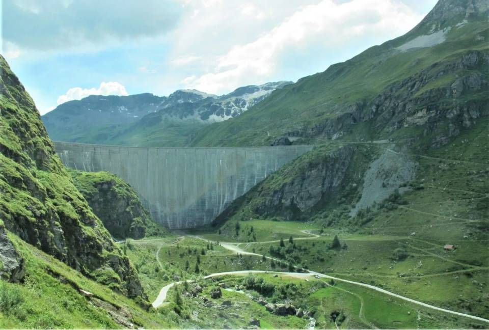 In einem üppig grünen Bergtal unter bewölktem Himmel erhebt sich ein Betonstaudamm; im Vordergrund verlaufen gewundene Feldwege.