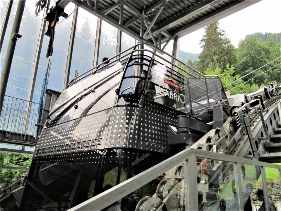 Nahaufnahme einer schwarzen Bergseilbahn, die an einer Station ankommt; ihre Metallkonstruktion ist vor einem grünen Wald deutlich zu erkennen.