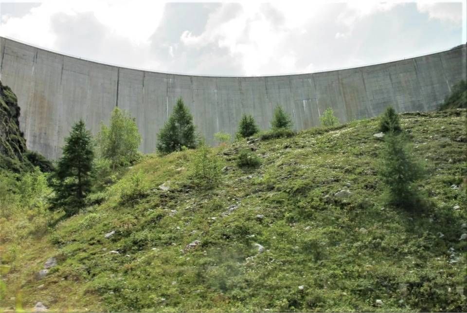 Unter einem bewölkten Himmel erhebt sich ein hoher, geschwungener Betonstaudamm über einem grasbewachsenen, mit Bäumen übersäten Hügel.