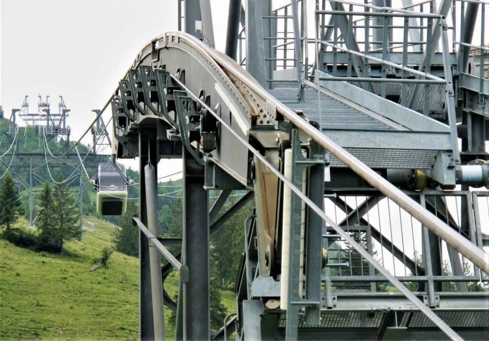 Eine Seilbahnstation mit einer gebogenen Metallschiene und einer im Hintergrund vor einem grünen Hügel sichtbaren Gondel an einem Seil.