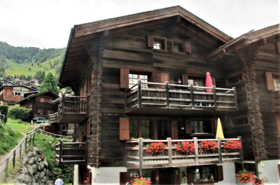 Ein rustikales, mehrstöckiges Holzchalet in einem Bergdorf mit zwei Balkonen, die mit roten Blumen geschmückt sind.