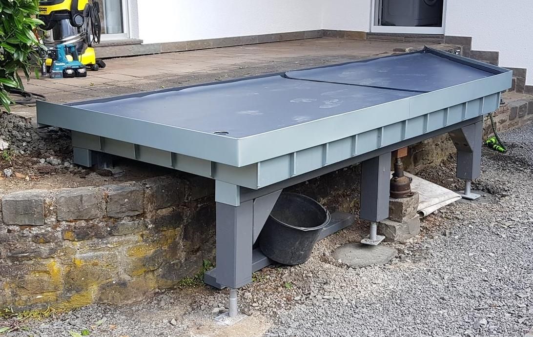 An der Seite eines Gebäudes steht neben einem Eimer eine Bank.