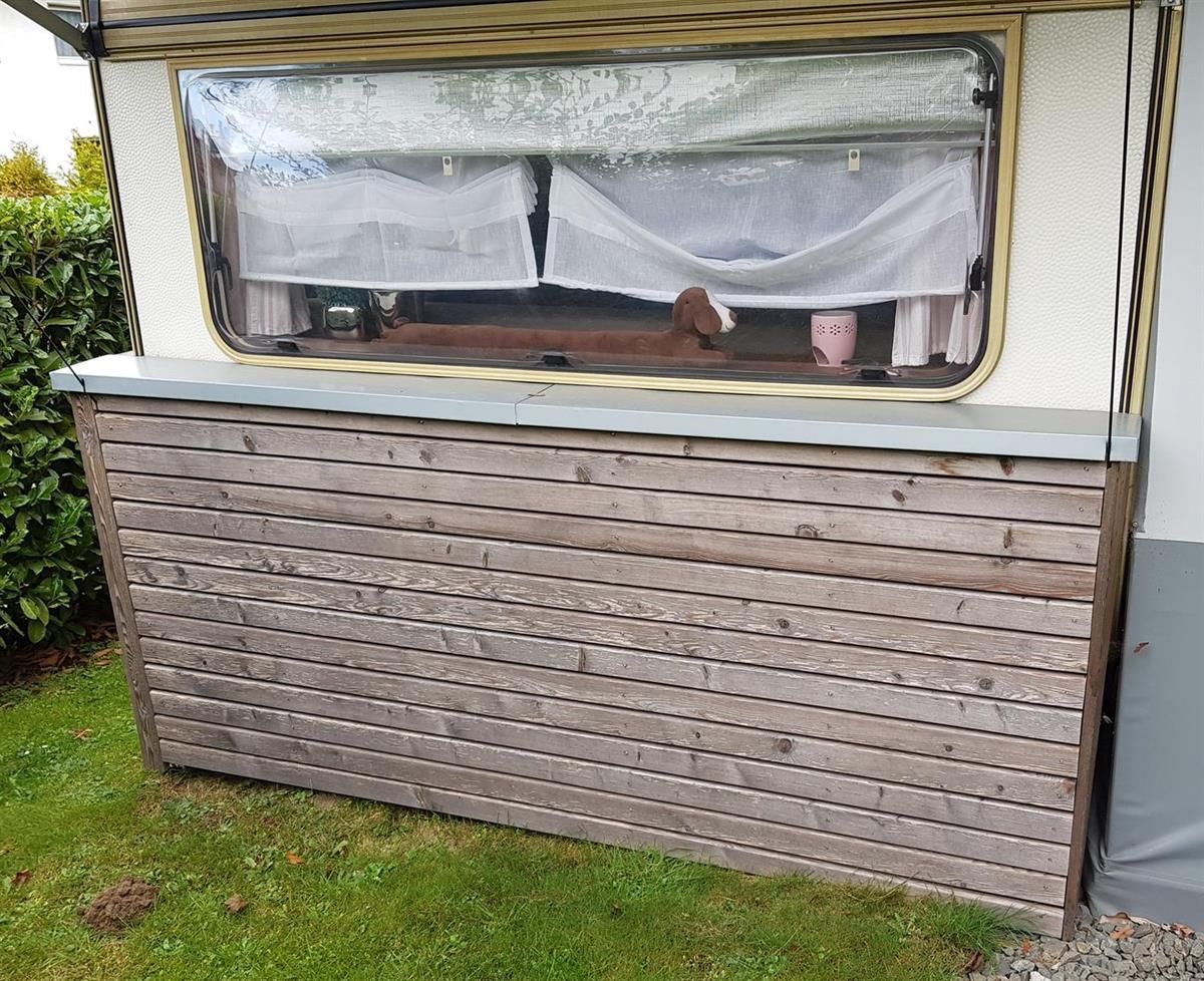 Ein Wohnwagen mit einer Holzwand und einem Fenster steht im Gras.