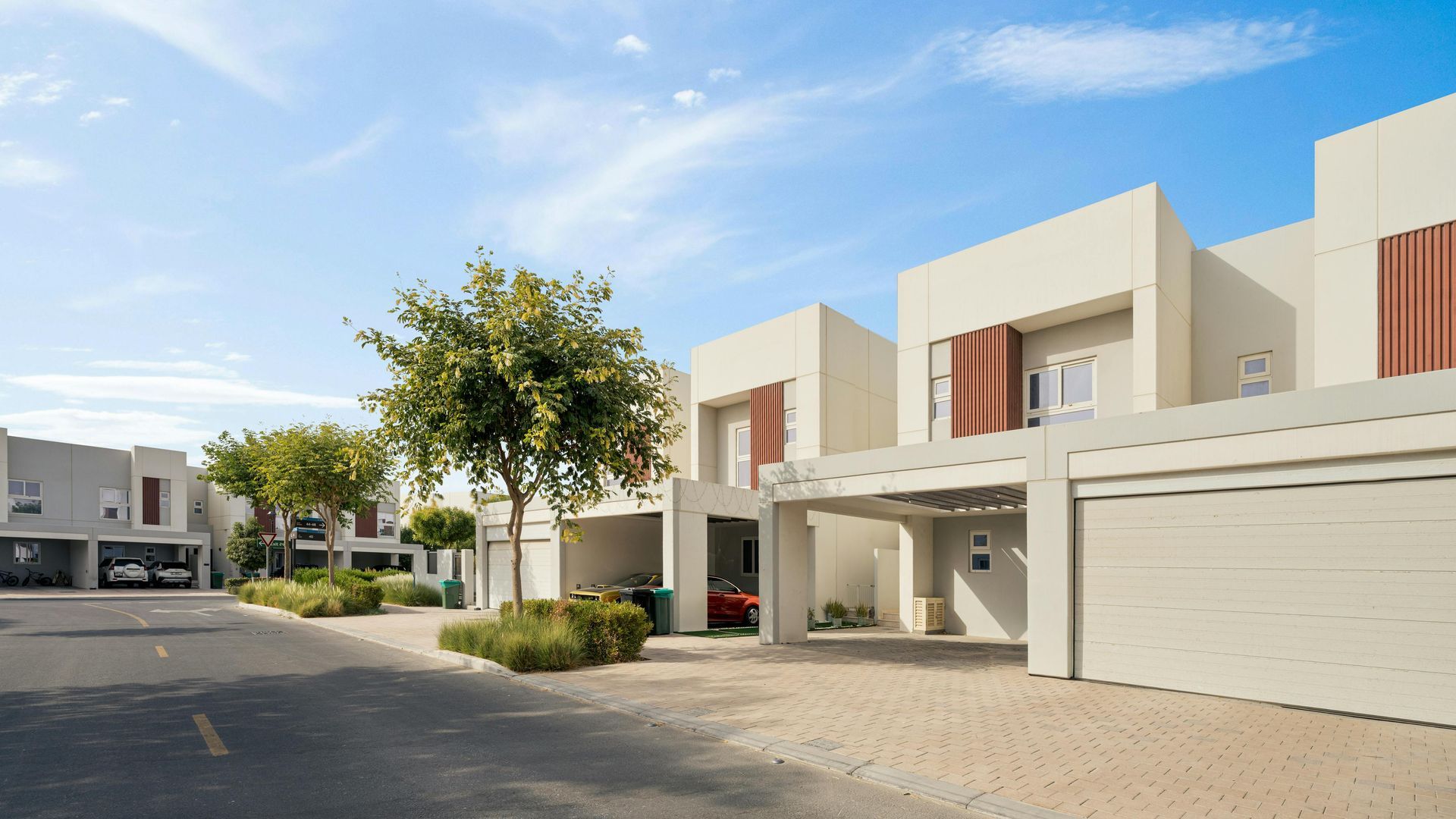 Modernos apartamentos de color beige con garajes bordean una tranquila calle residencial bajo un cielo azul.
