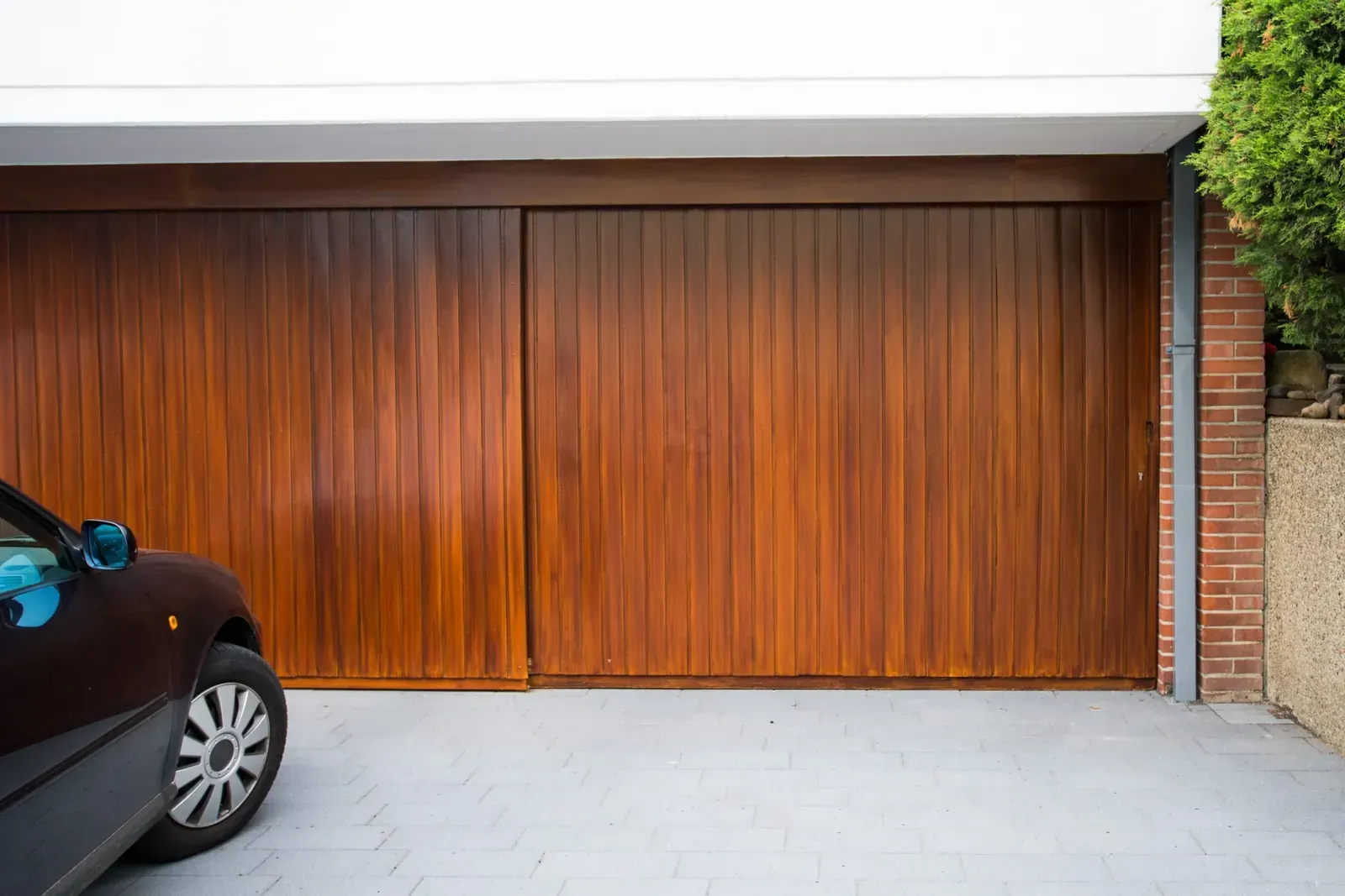 Puertas de garaje dobles de madera junto a un coche aparcado y una pared de ladrillos.