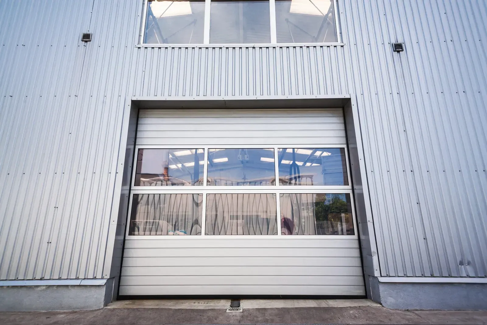 Fachada de un edificio industrial con una gran puerta de garaje blanca y ventanas que reflejan el cielo.