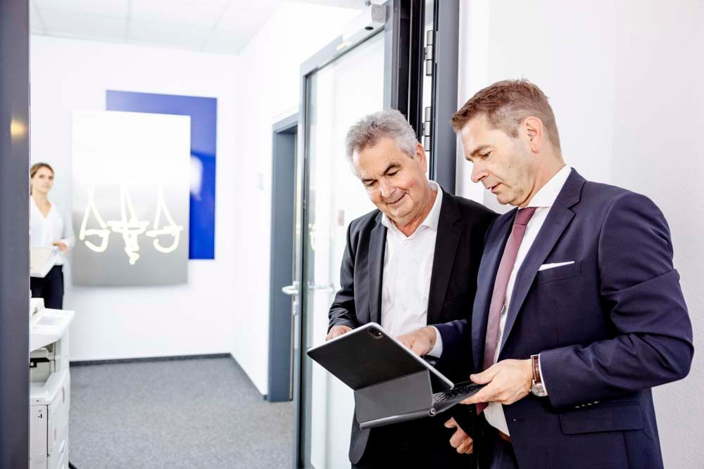 Zwei Männer in Anzügen schauen in einem Büroflur auf ein Tablet; eine Frau steht daneben.