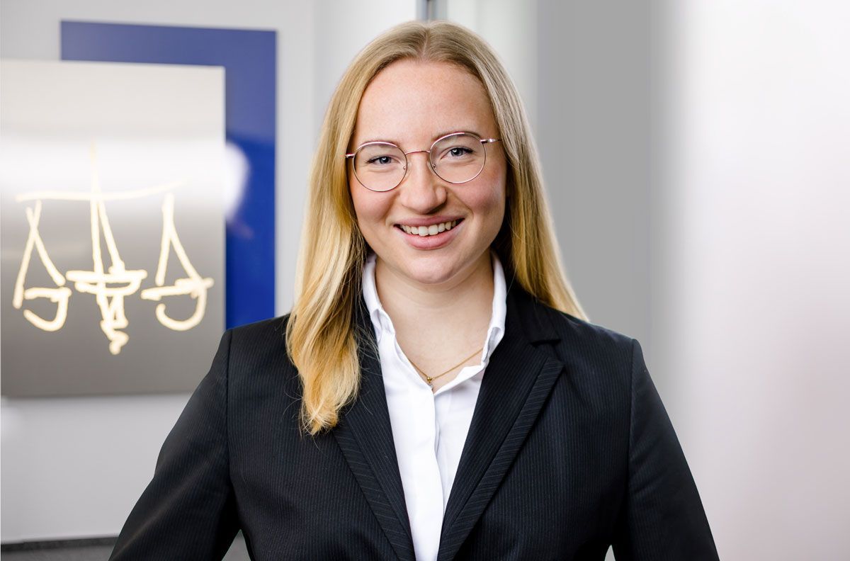 Eine Frau im Anzug und mit Brille lächelt vor einem Schild mit der Aufschrift „Aaa“.