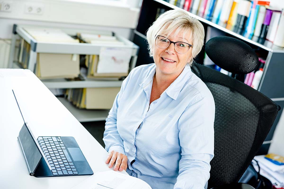 Eine Frau mit Brille lächelt, während sie an einem Schreibtisch sitzt; im Hintergrund sind ein Tablet und Aktenregale zu sehen.