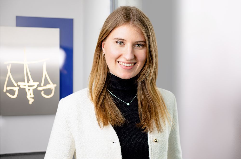 Eine Frau im Anzug und mit Brille lächelt vor einem Schild mit der Aufschrift „Aaa“.