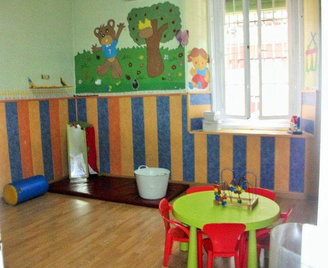 Una colorida sala de juegos preescolar con una mesa redonda verde, sillas rojas, una alfombra, un cubo blanco y un cuadro de osos en la pared.