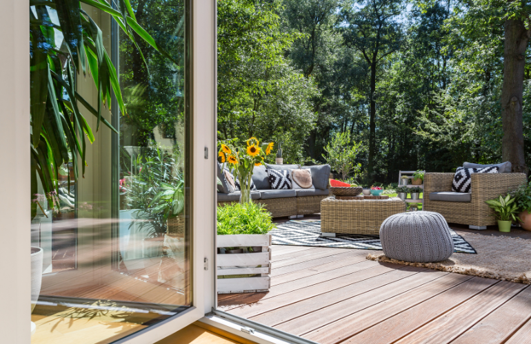 Terrasse extérieure avec porte vitrée ouverte, meubles en osier, plantes et soleil.