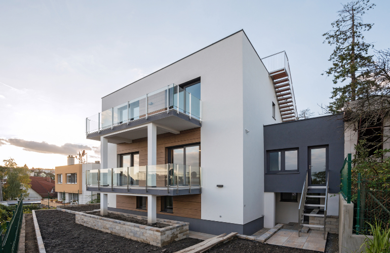 Bâtiment moderne blanc à plusieurs étages avec balcons en verre et escalier extérieur à flanc de colline.