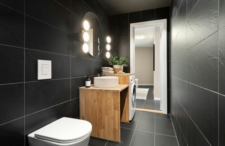 Salle de bain moderne avec murs carrelés noirs, vanité en bois et vue sur une autre pièce.