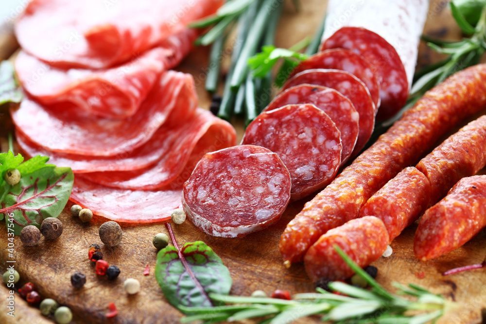 Salami- und Wurstscheiben auf einem Holzbrett mit Kräutern und Pfefferkörnern.