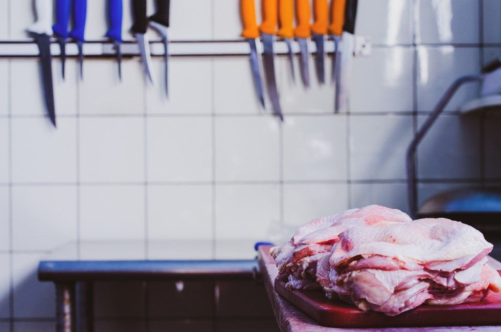 Rohes Hühnerfleisch auf einem Schneidebrett in einer Küche, an deren Wand Messer hängen.
