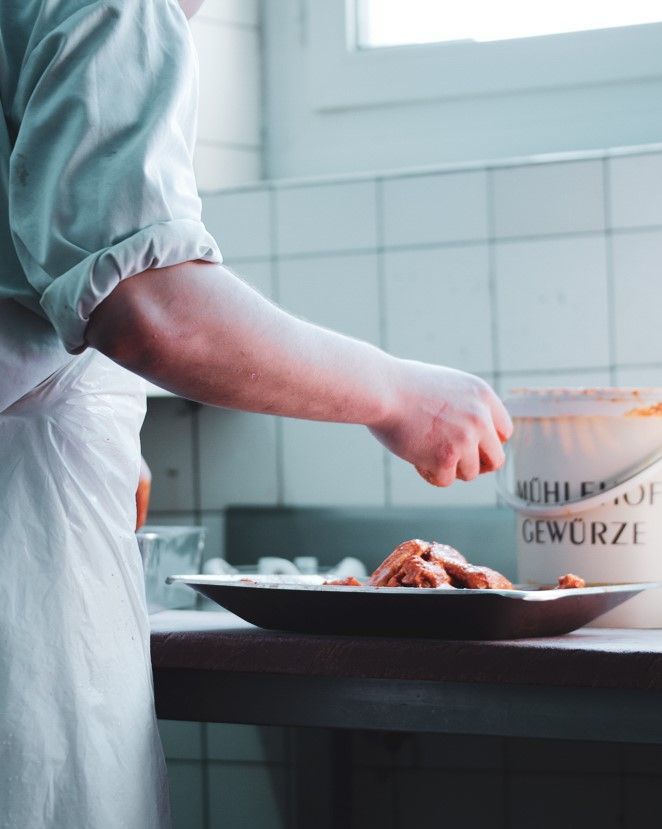 Der Koch bestreut Speisen auf einem Teller in einer Küche mit weissen Fliesenwänden mit Gewürzen.