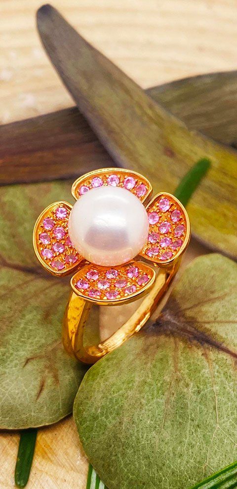 Bague en or avec un centre en perle et un motif de fleur en pierre précieuse rose sur des feuilles vertes.