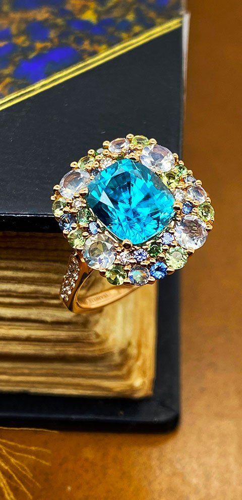 Une bague en or avec une grosse pierre centrale bleue et un halo de plus petites pierres précieuses colorées.