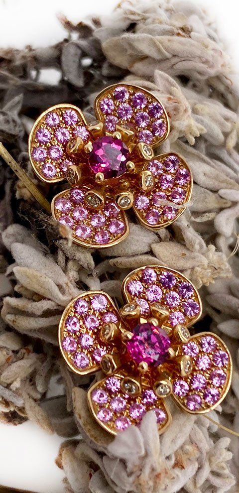 Boucles d'oreilles florales en or avec pierres précieuses roses sur un lit de plantes séchées.