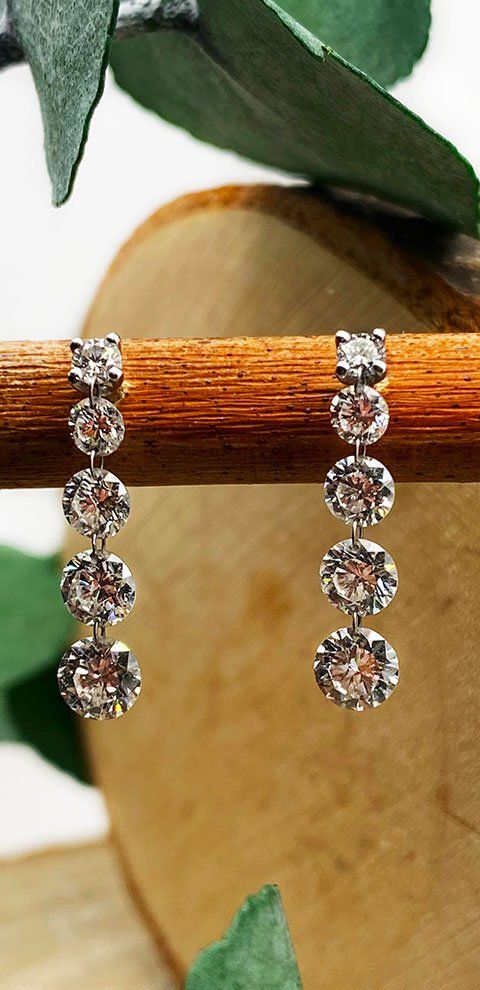 Deux boucles d'oreilles pendantes en diamant sont suspendues à un goujon en bois, avec de la verdure en arrière-plan.