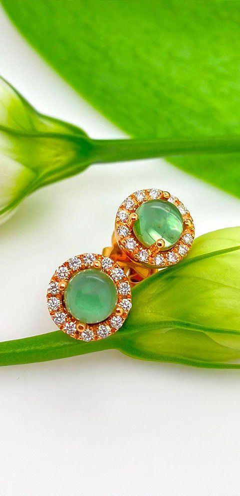 Boucles d'oreilles en or avec pierres vertes, entourées de diamants, reposent sur un fond vert feuille et blanc.