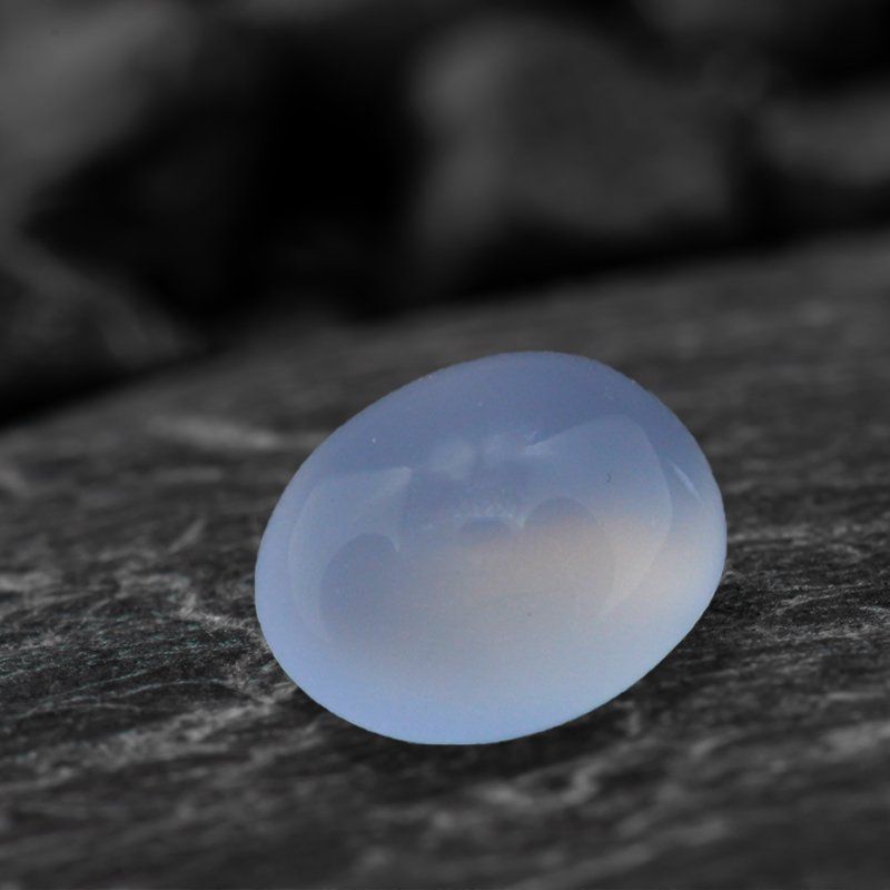 Calcédoine de forme ovale reposant sur une surface sombre et texturée, arrière-plan flou.