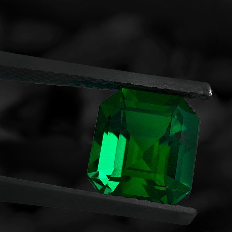 Pierre précieuse émeraude tenue par une pince, couleur verte, taille rectangulaire, fond sombre.