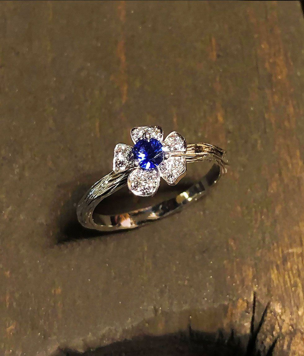 Bague avec une pierre centrale en saphir bleu entourée de diamants blancs dans un sertissage fleur, sur une surface texturée.