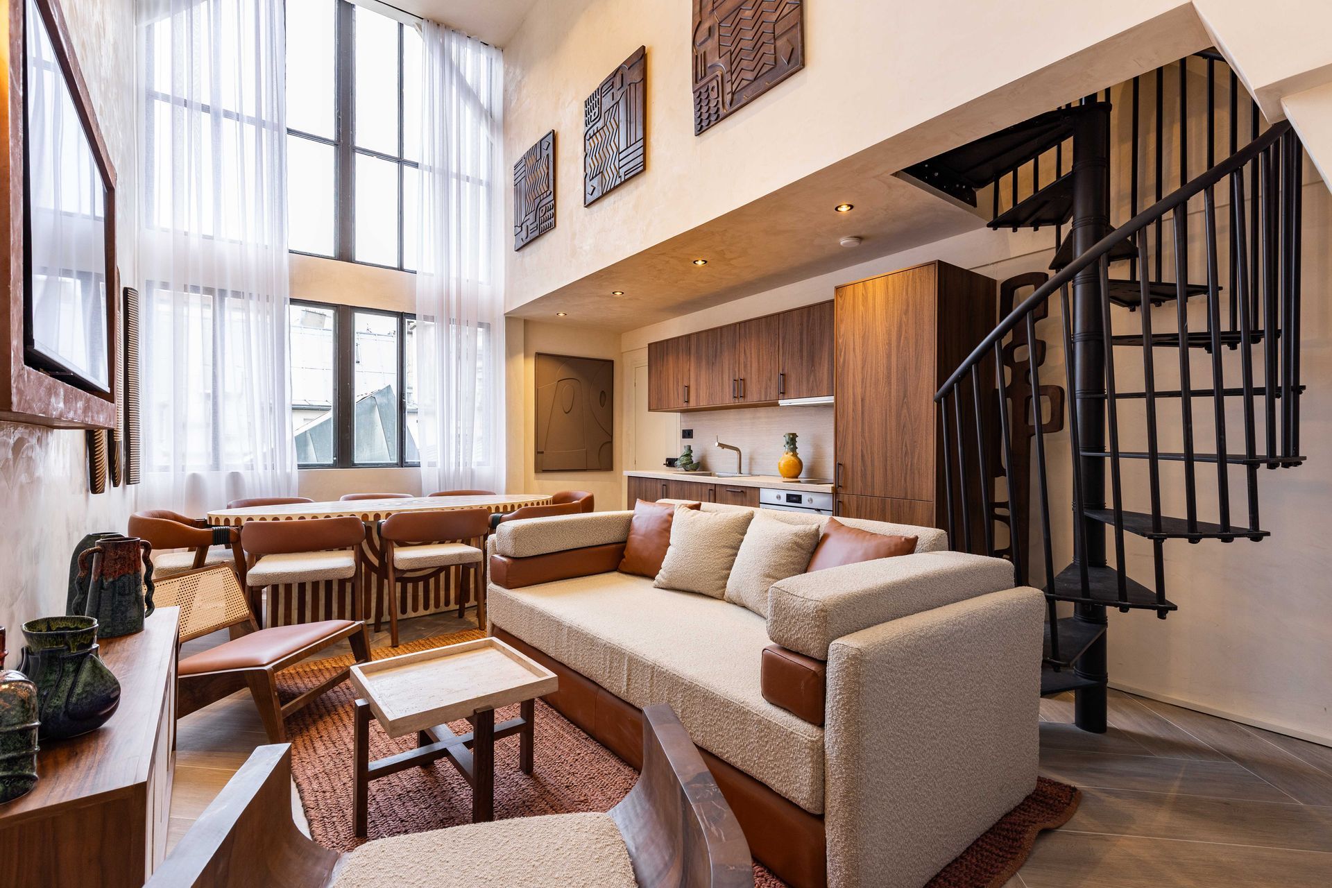 Loft apartment with living area, kitchen, and spiral staircase. Tall windows, neutral colors, and decorative art.