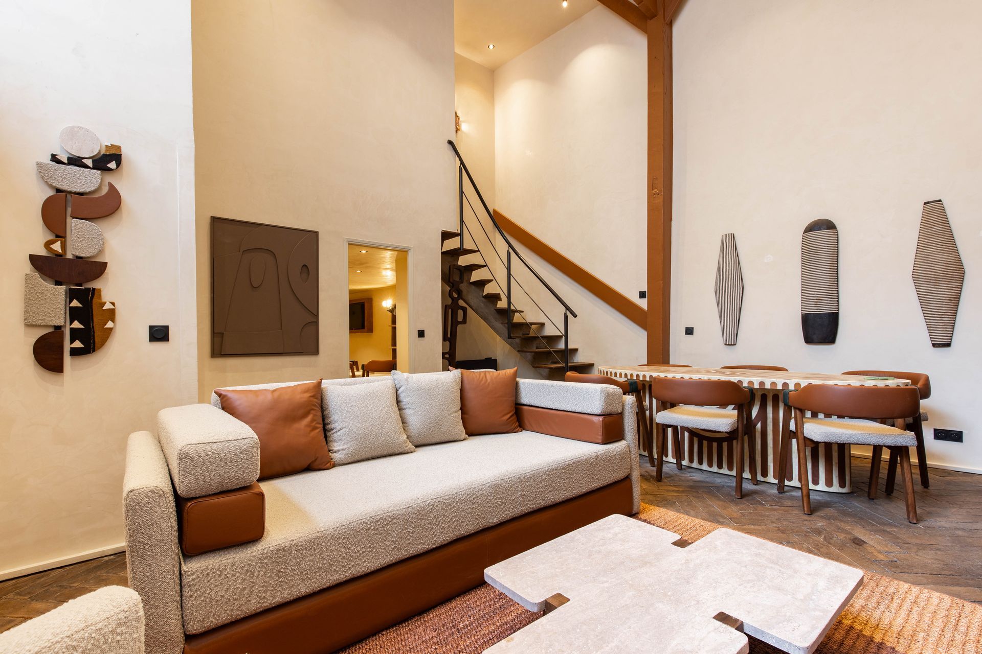 Spacious living room with sofa, dining table, and staircase. Beige walls, wood accents, decorative art.