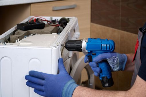 Une personne utilise une perceuse bleue pour réparer le côté d'une machine à laver blanche.