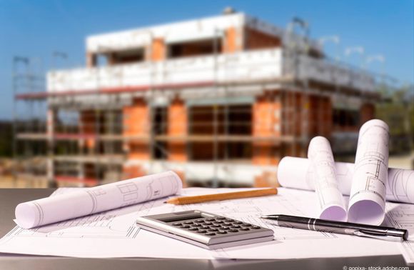 Ein Tisch mit Grundrissplänen, einem Taschenrechner und einem Stift leigen vor einem Neubau