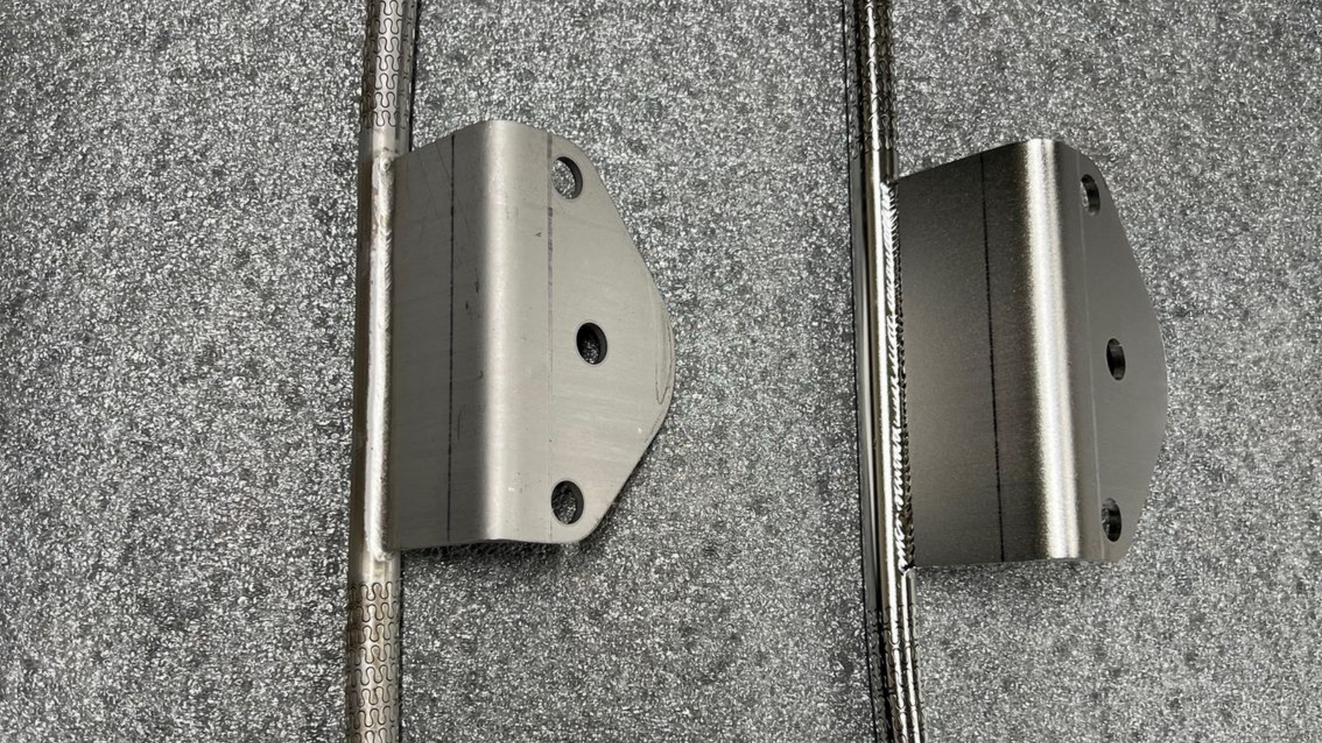 Deux supports métalliques gris, chacun comportant trois trous, sur une surface texturée avec des lignes.