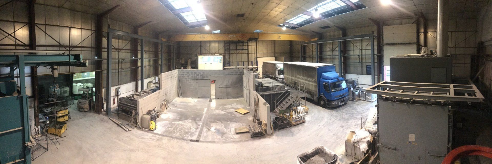 À l'intérieur d'un grand atelier industriel. Un camion bleu est stationné. D'autres machines et équipements sont présents.