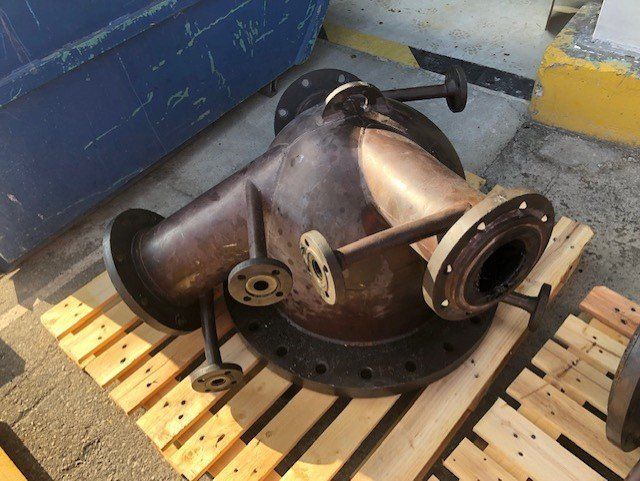 Gros tuyau en métal sombre en forme de Y avec plusieurs raccords à brides, posé sur une palette en bois à l'extérieur.