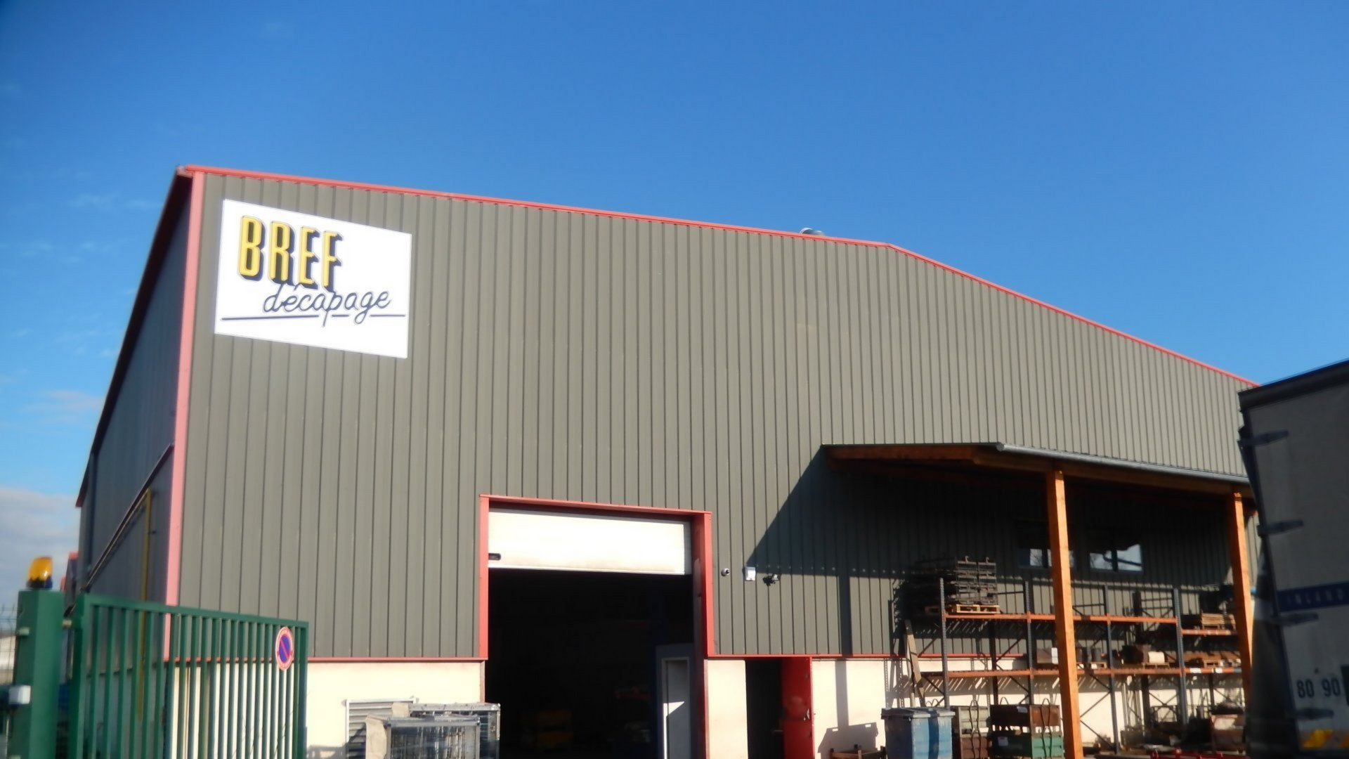Bâtiment industriel gris avec des bordures rouges et une enseigne d'entreprise sous un ciel bleu.