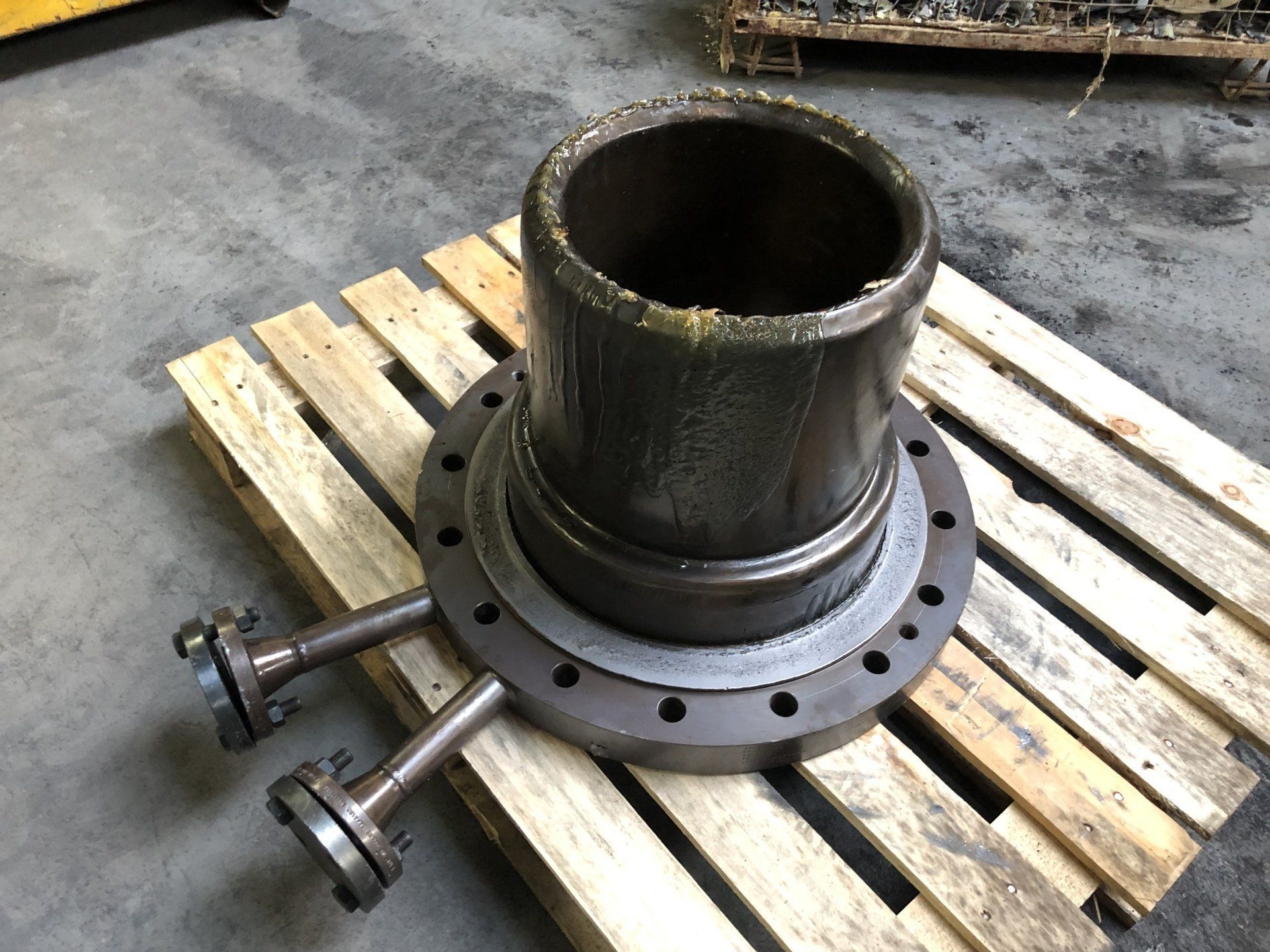 Composant métallique industriel avec corps cylindrique, base à bride et deux tuyaux fixés, reposant sur une palette en bois.