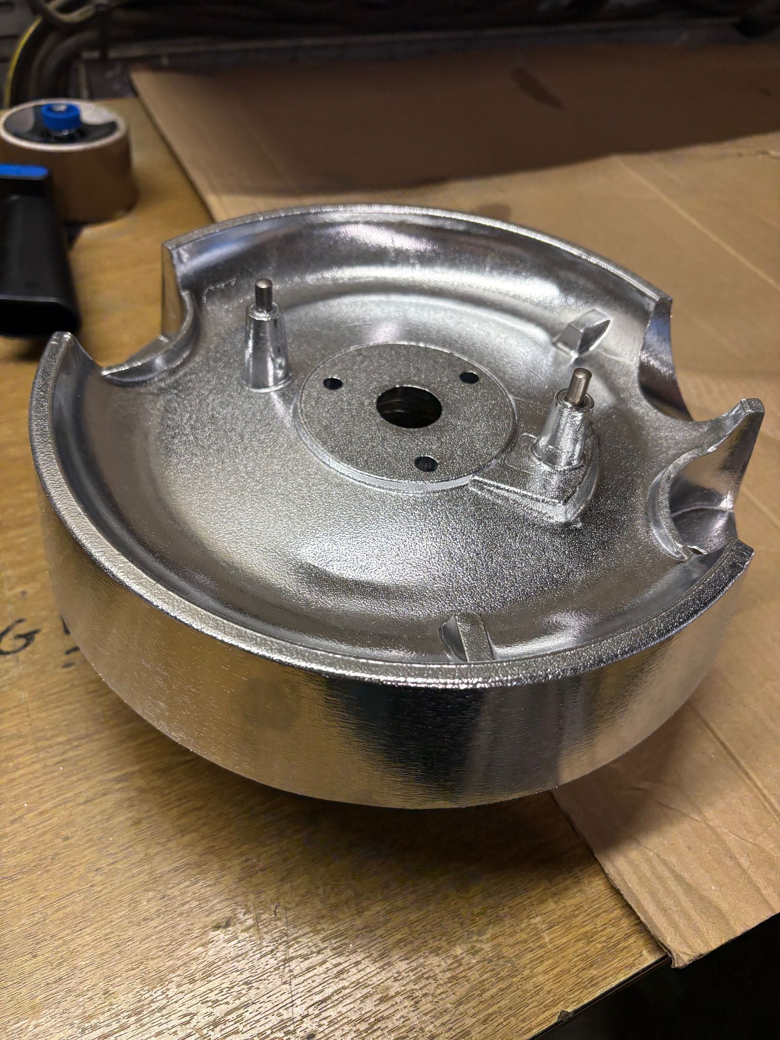 Un élément métallique circulaire, brillant et argenté, muni d'un trou central et de languettes en relief, repose sur une surface en carton.