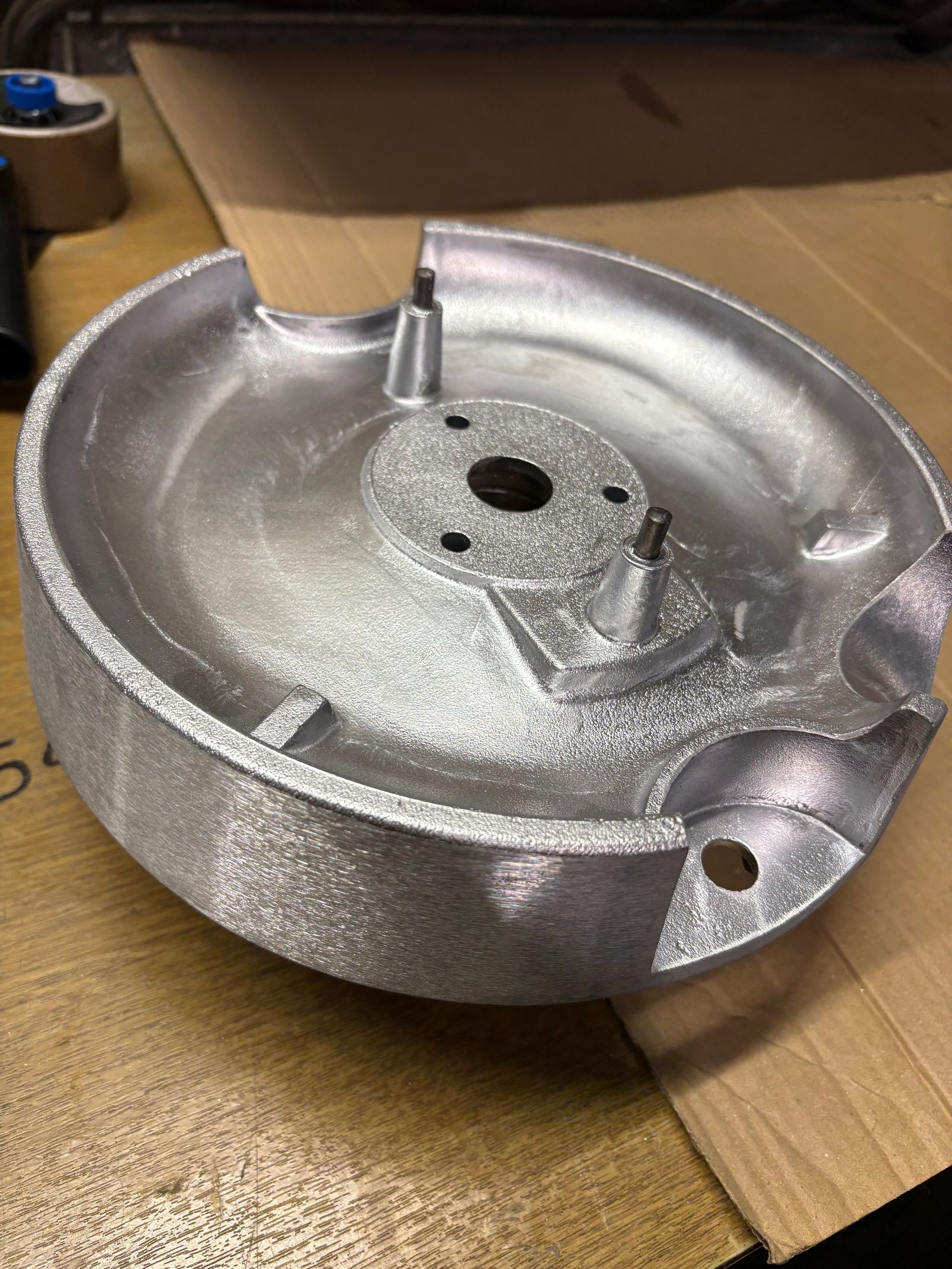 Pièce circulaire en métal coulé, non finie, avec un moyeu central, deux petits goujons et des bords crantés, sur une surface en carton.