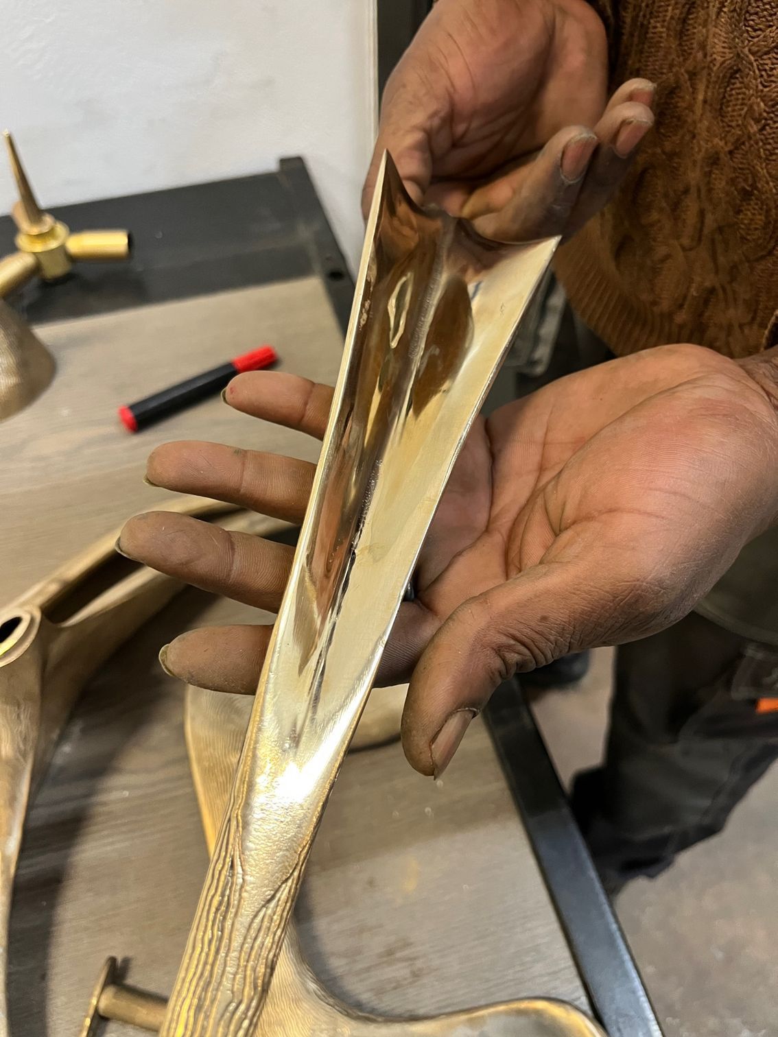 Des mains tiennent et présentent une pièce métallique incurvée de couleur dorée avec un intérieur réfléchissant ; décor d'atelier.