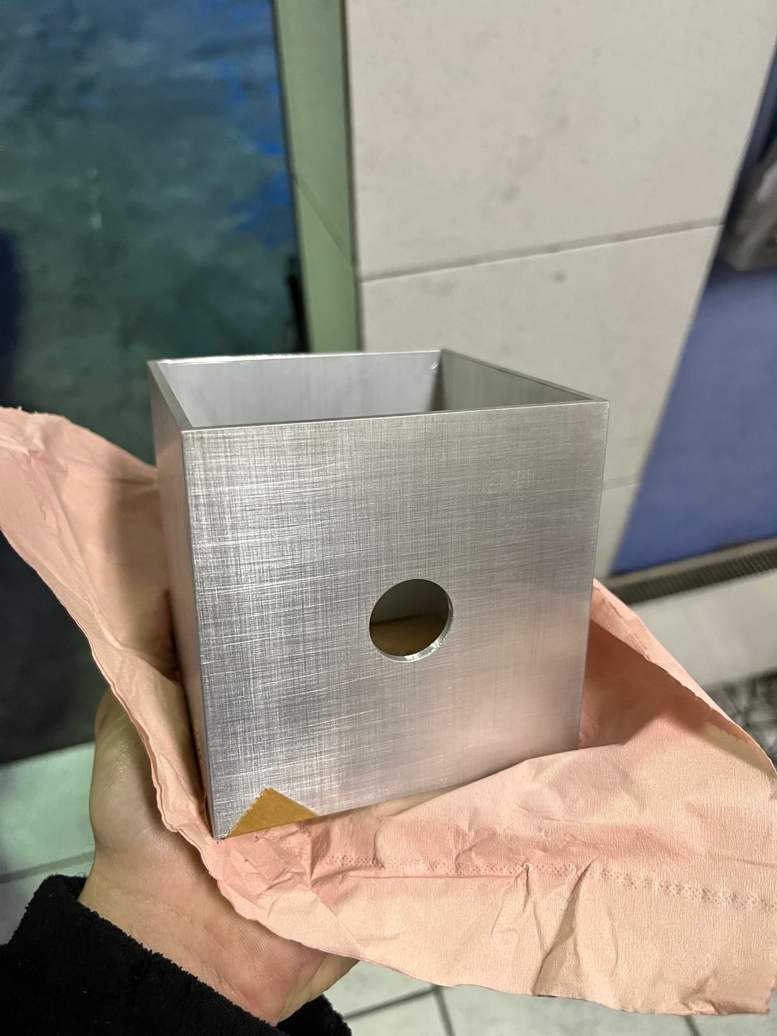Personne tenant un récipient métallique carré percé d'un trou sur un côté ; posé sur un tissu rose.