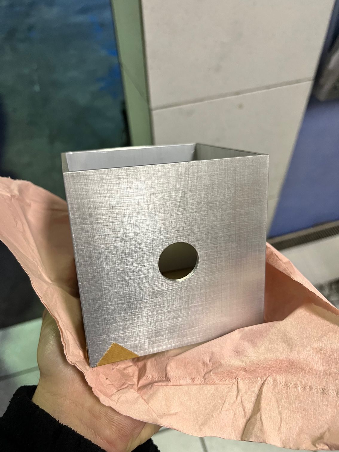 Une main tient une boîte carrée en métal brossé, percée d'un unique trou circulaire au centre, posée sur un tissu froissé couleur pêche.