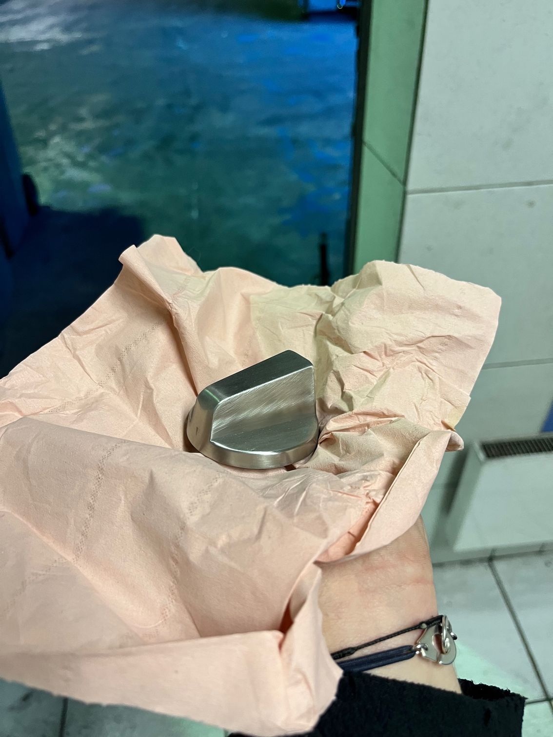 Main tenant un objet métallique argenté et angulaire enveloppé dans du papier de soie rose ; eau en arrière-plan.